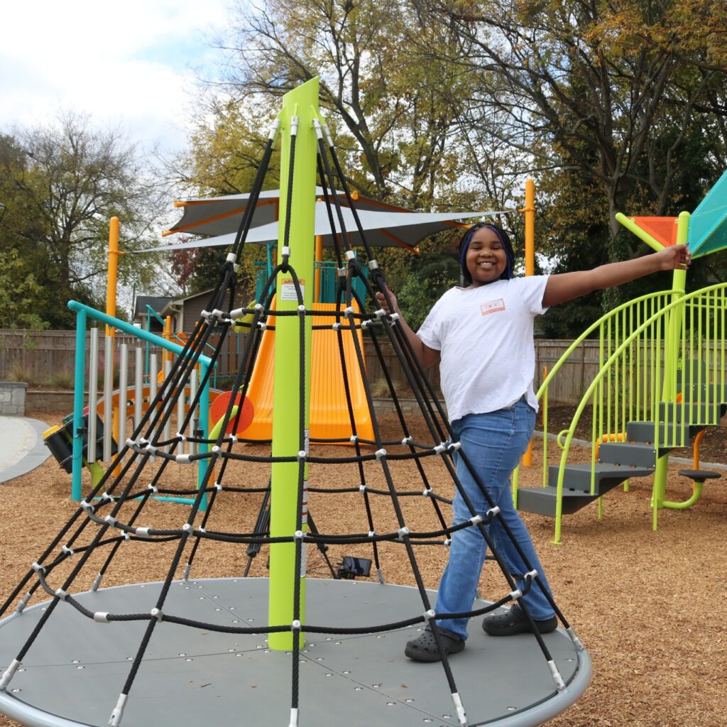 A smiling girl with blue-tipped braids stands on a round play structure with black rope netting, beside bright lime-green and orange playground equipment.